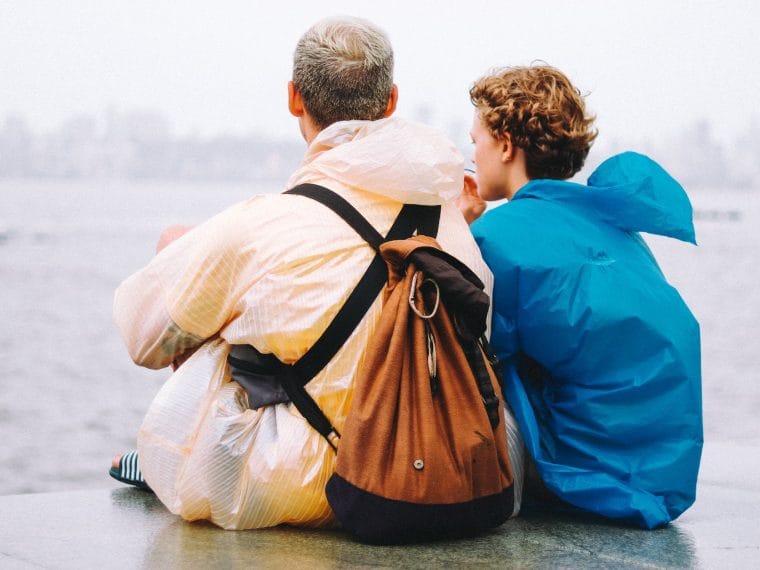 man and teenage boy in raincoats sitting by a sea