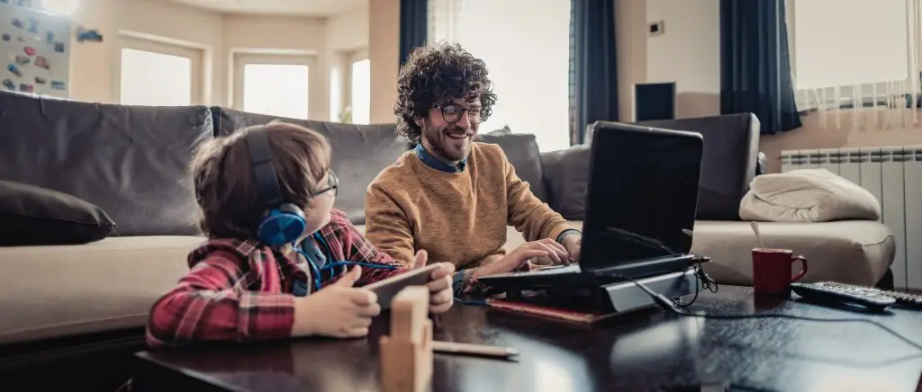 Father working on a laptop next to his son who is wearing headphones and playing on a tablet.  This photo highlights the need for self-compassion for parents